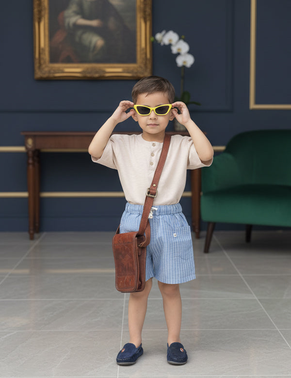 Stylish young boy wearing yellow sunglasses, beige shirt, blue striped shorts, and brown leather bag indoors