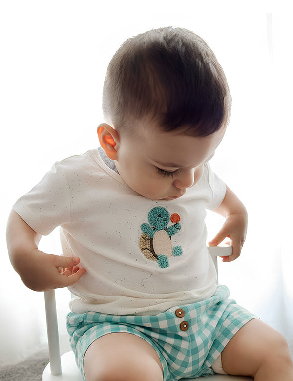 Toddler boy sitting on a white chair wearing a white t-shirt with a turtle graphic and blue checkered shorts