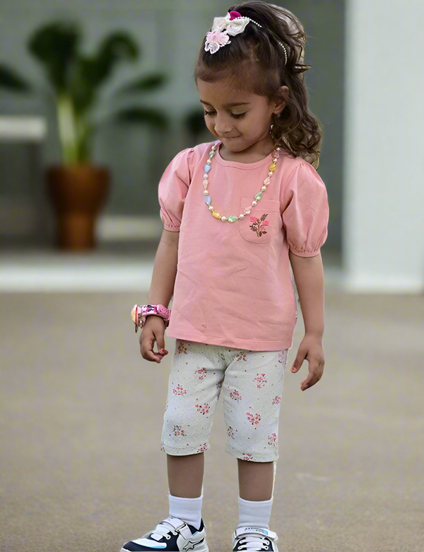 Adorable young girl, appearing to be of South Asian descent, wearing a salmon pink Oh My Baby organic cotton top with puff sleeves and a floral embroidered pocket, paired with grey floral print capri leggings. Shop cute kids' outfits online in India.