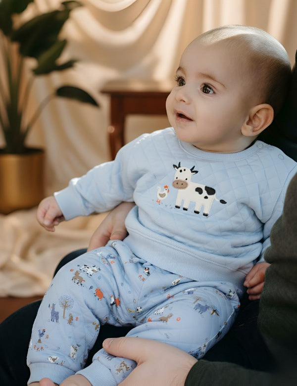Smiling baby wearing blue organic quilted sweatshirt and farm-print pants — cozy baby winter clothes outfit.