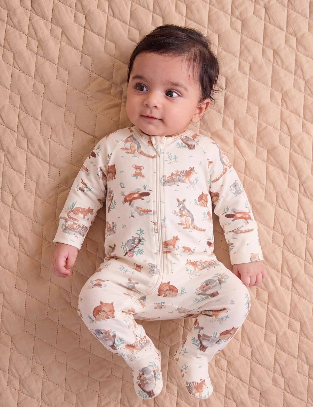 Baby model lying on a quilted tan blanket wearing an organic cotton footie romper featuring an all-over Australian animal print.