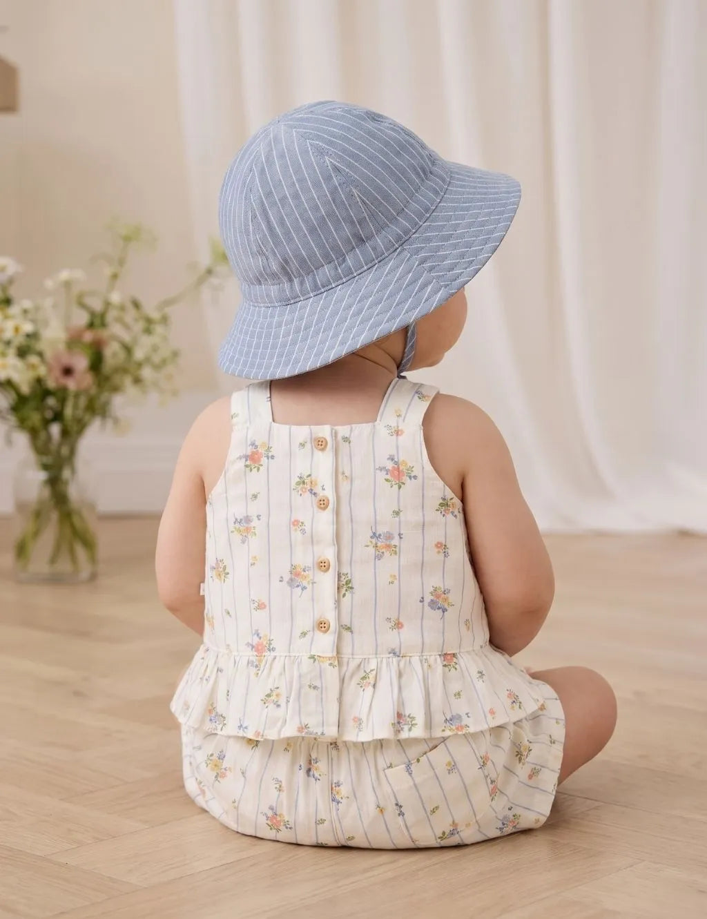 Back view of a baby sitting on a wooden floor wearing an organic cotton blue striped floral peplum top with back buttons, matching shorts, and a bucket hat.