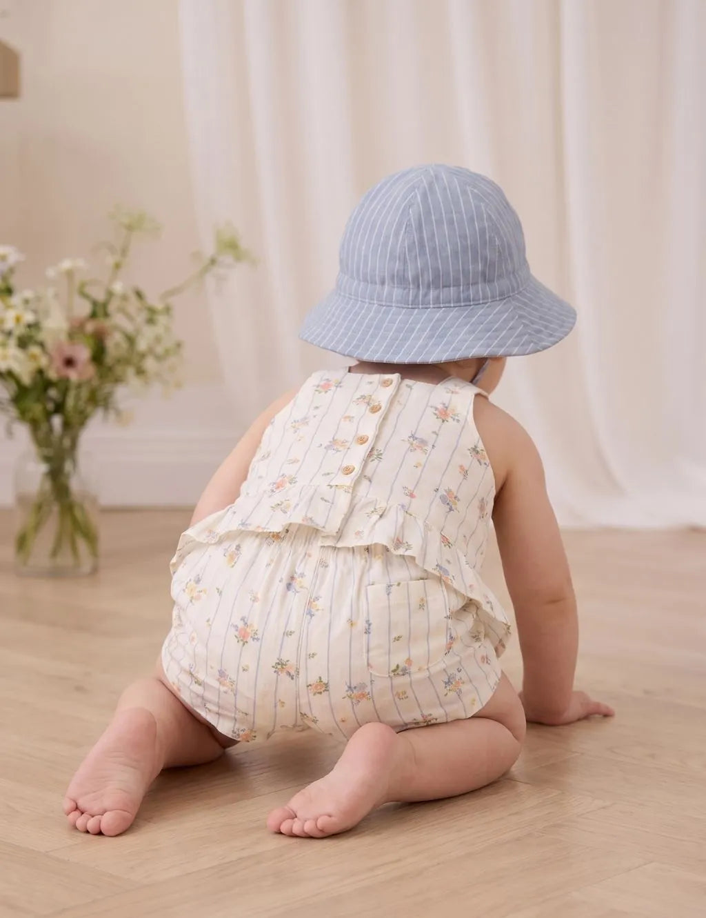 Back view of a baby crawling on a wooden floor wearing an organic cotton blue striped floral peplum top and matching shorts with a bucket hat.
