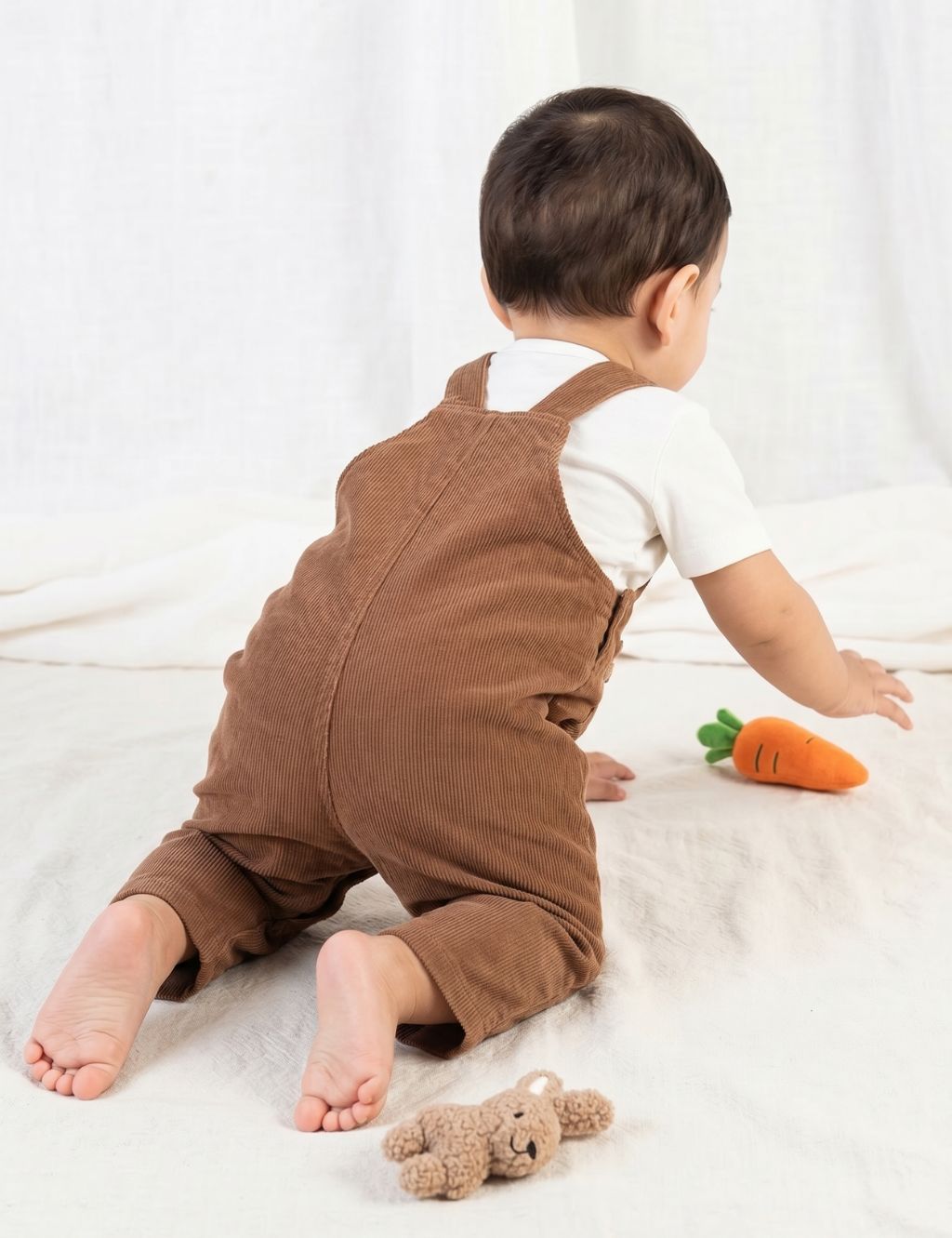 Back view of a baby wearing an organic cotton brown corduroy jumpsuit and crawling toward a toy carrot.