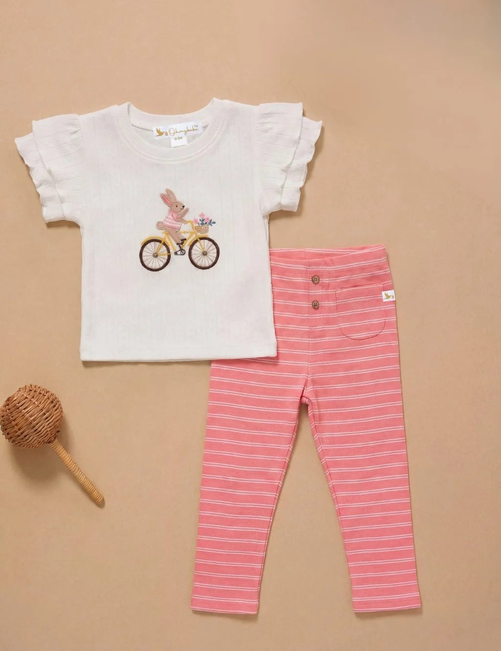 Flatlay of an organic cotton white ruffled-sleeve top with an embroidered bunny on a bicycle paired with pink striped leggings and a small woven rattle.