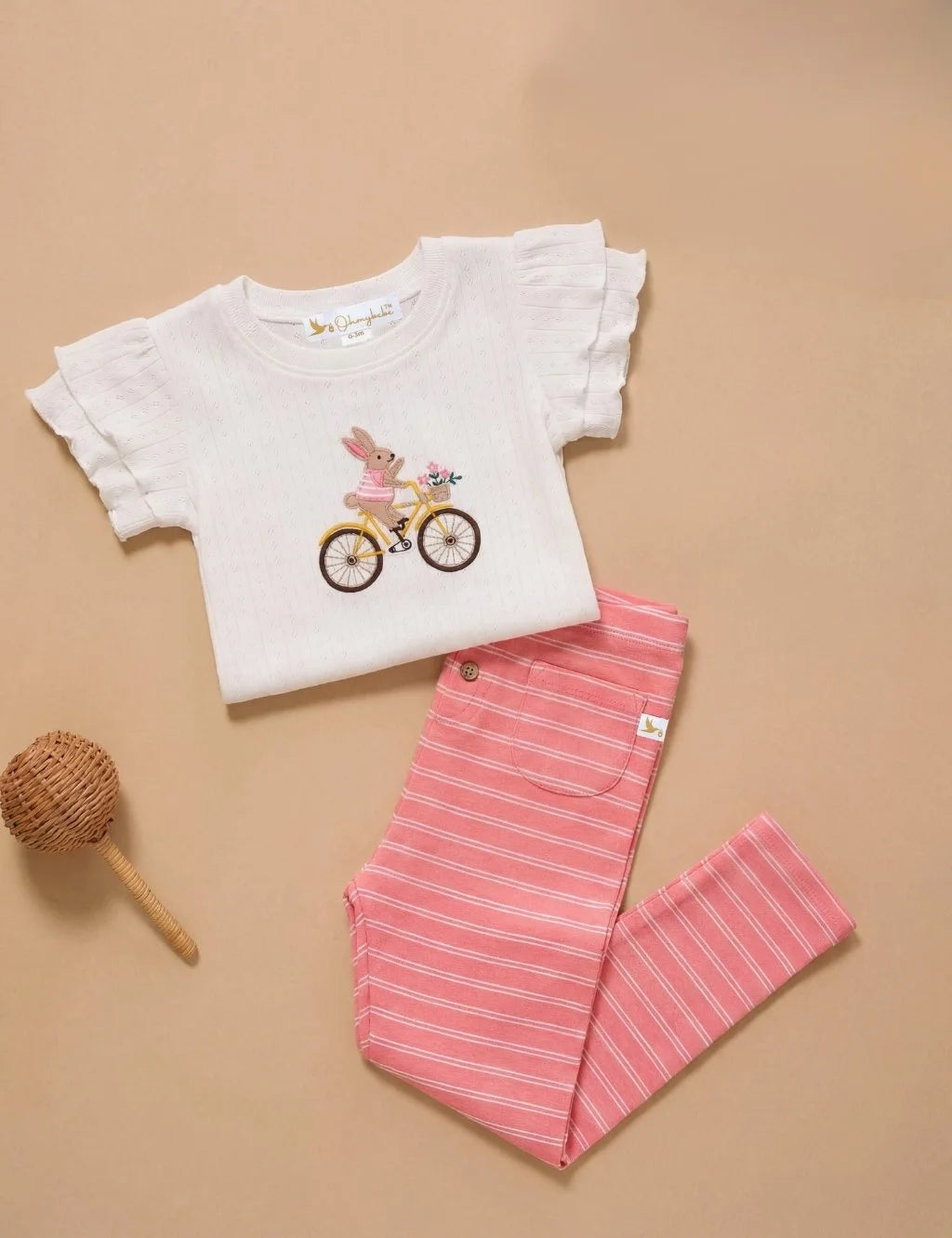 Flatlay of an organic cotton white ruffled-sleeve top with an embroidered bunny on a bicycle paired with pink striped leggings and a small woven rattle