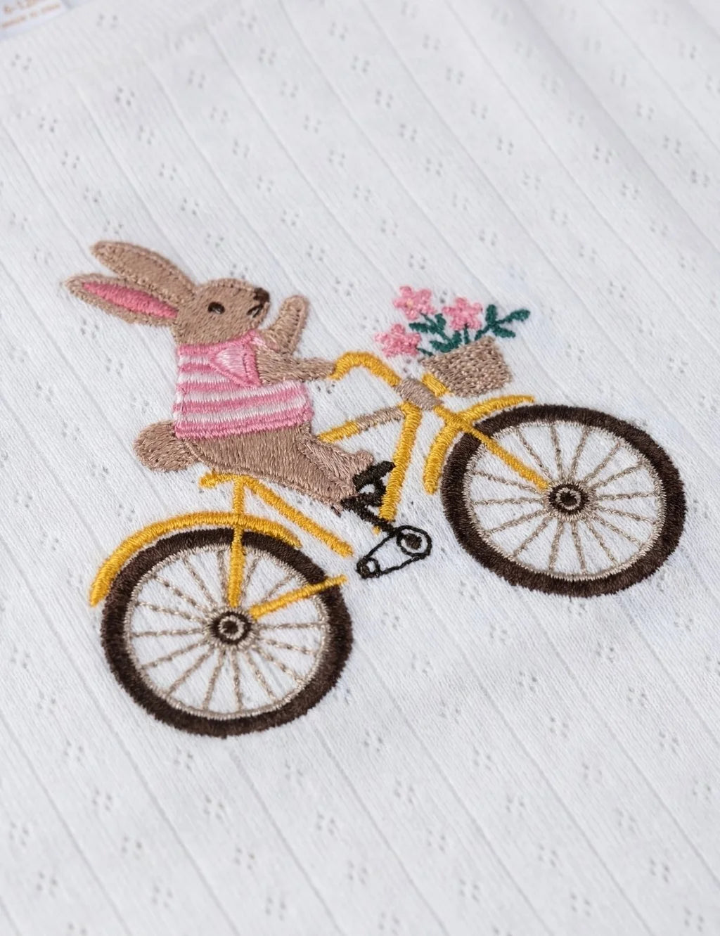 Close-up flatlay of an organic cotton white fabric featuring detailed embroidery of a bunny wearing a striped shirt on a yellow bicycle with a basket of pink flowers