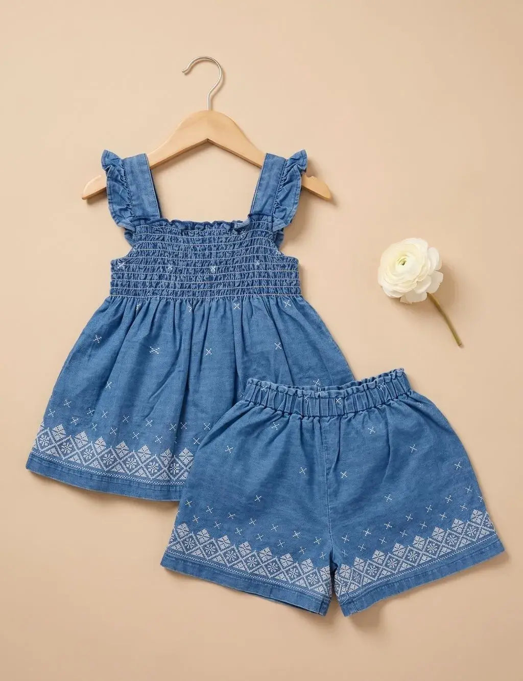 Flatlay of an organic cotton denim blue smocked top and shorts set with white embroidery next to a white flower.