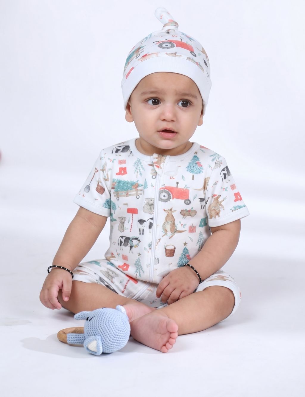Baby sitting on the floor wearing an organic cotton farm print shortie onesie with a matching knotted hat.