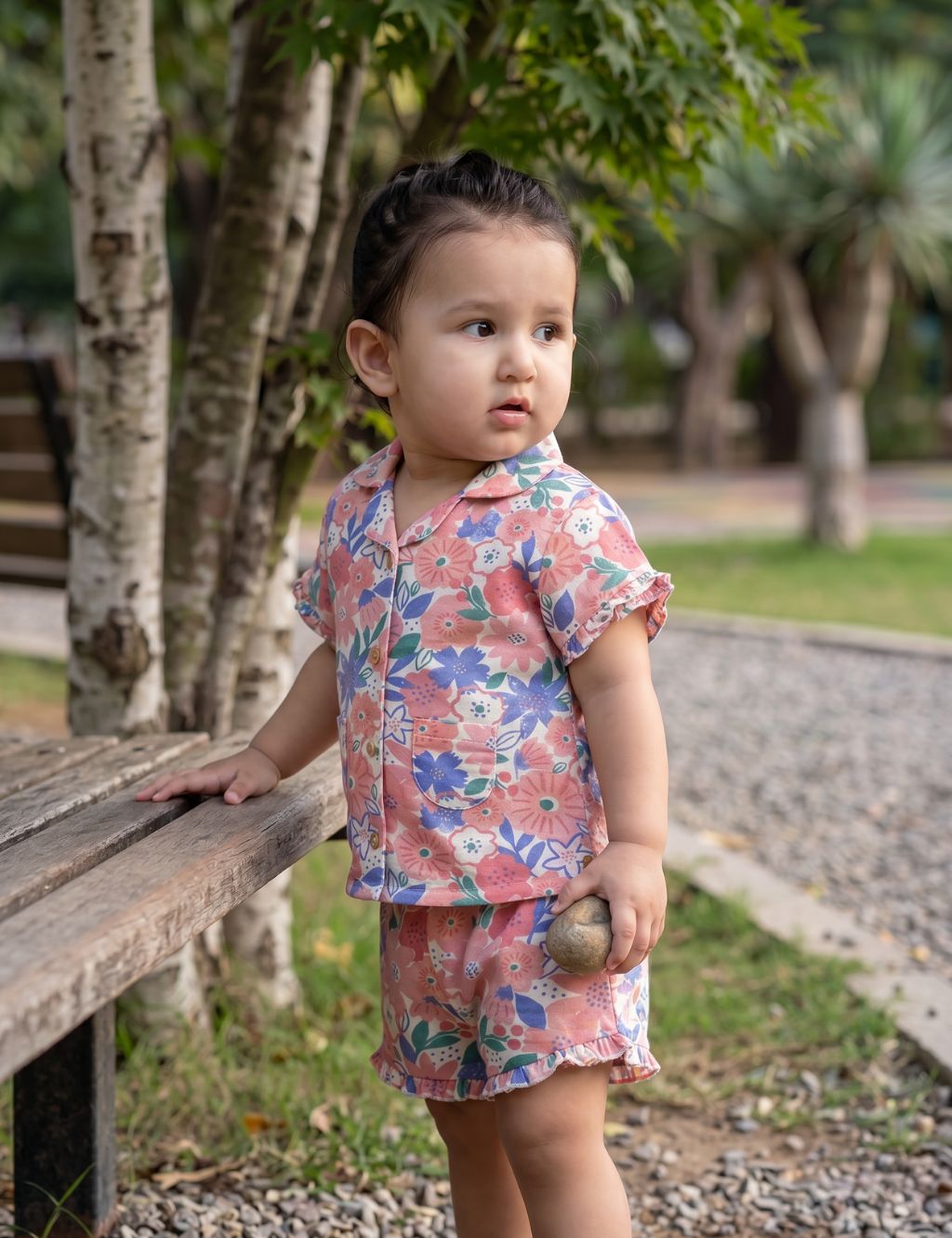Little toddler girl wearing an organic cotton pink floral print short-sleeve button-down shirt and matching shorts outdoors by a wooden bench