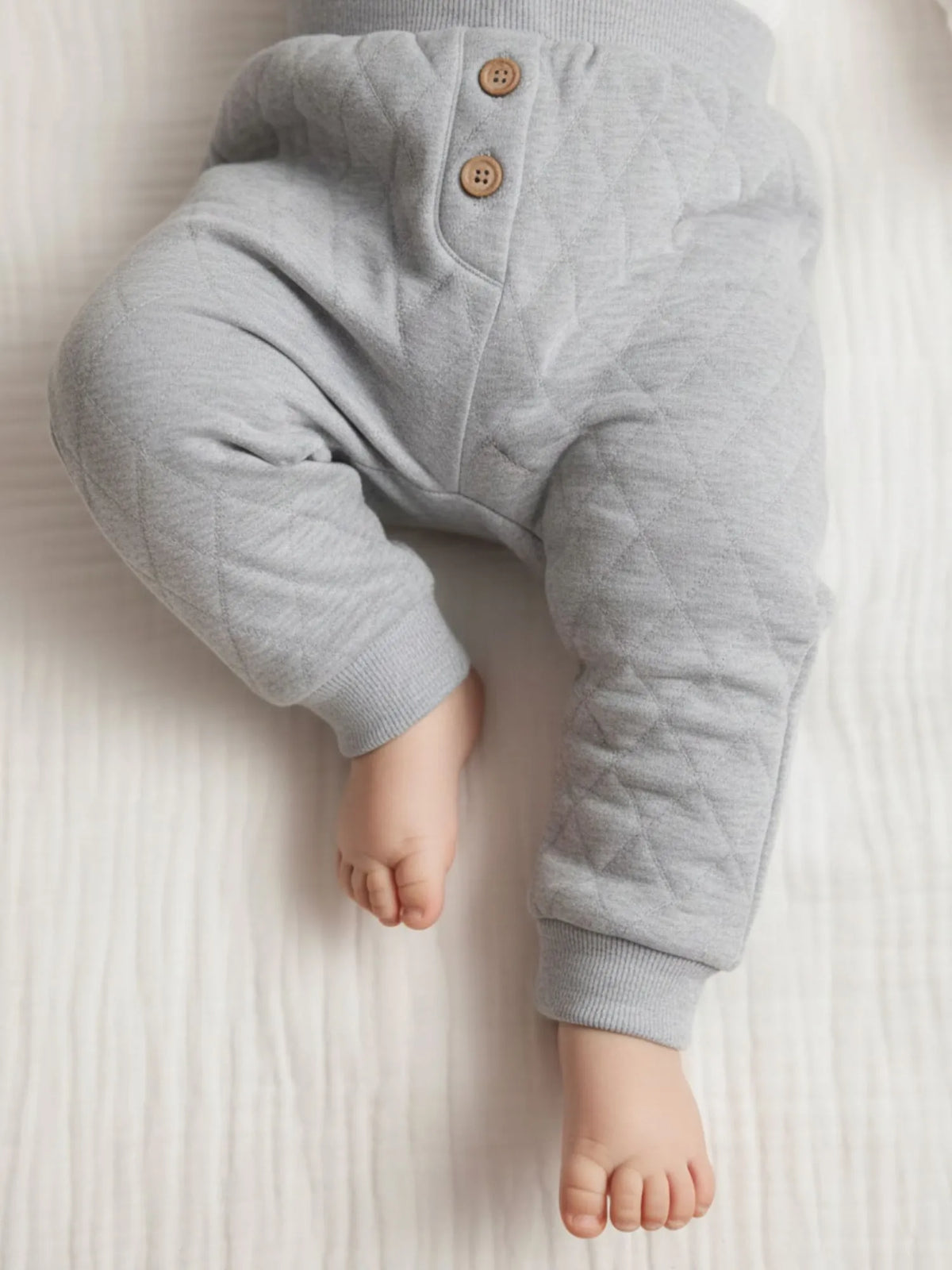 Baby lying down wearing organic cotton grey quilted pants with decorative wooden buttons