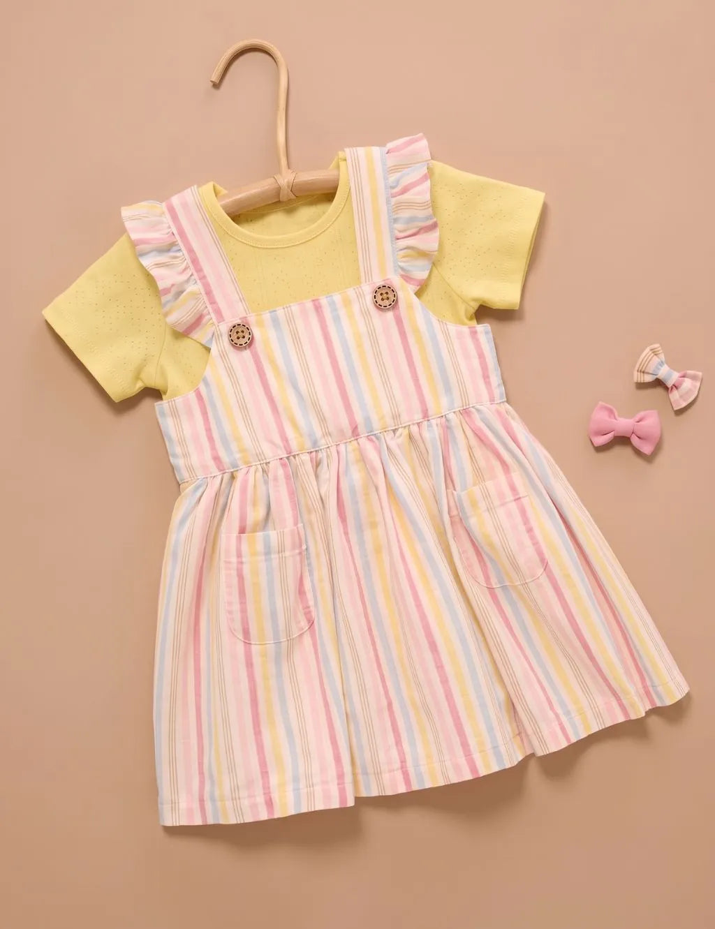 Flatlay of an organic cotton pastel striped pinafore dress with pockets and a yellow t-shirt set next to two hair bows