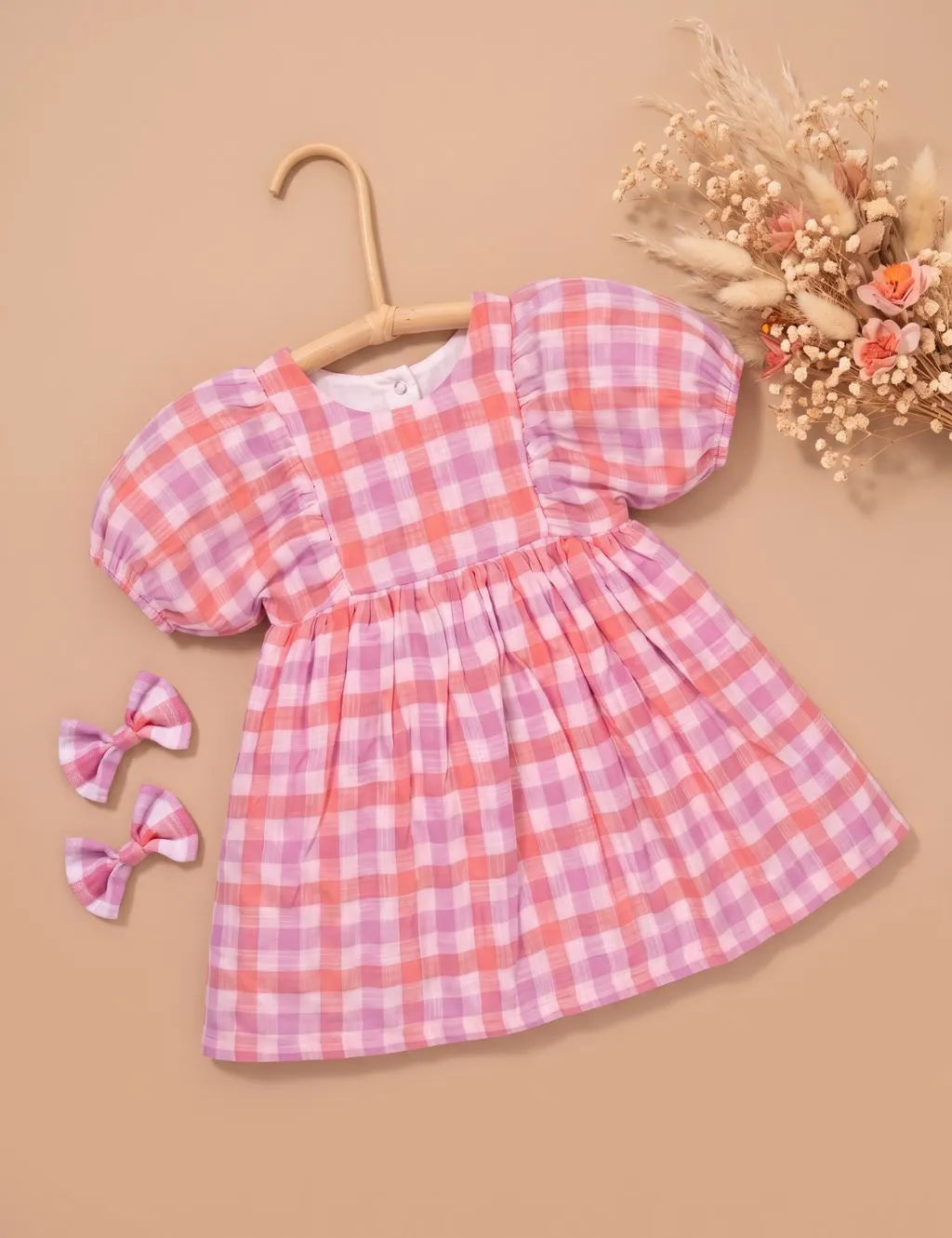 Hanging display of an organic cotton pink and purple checkered short-sleeve dress paired with matching hair bows and dried floral decor.