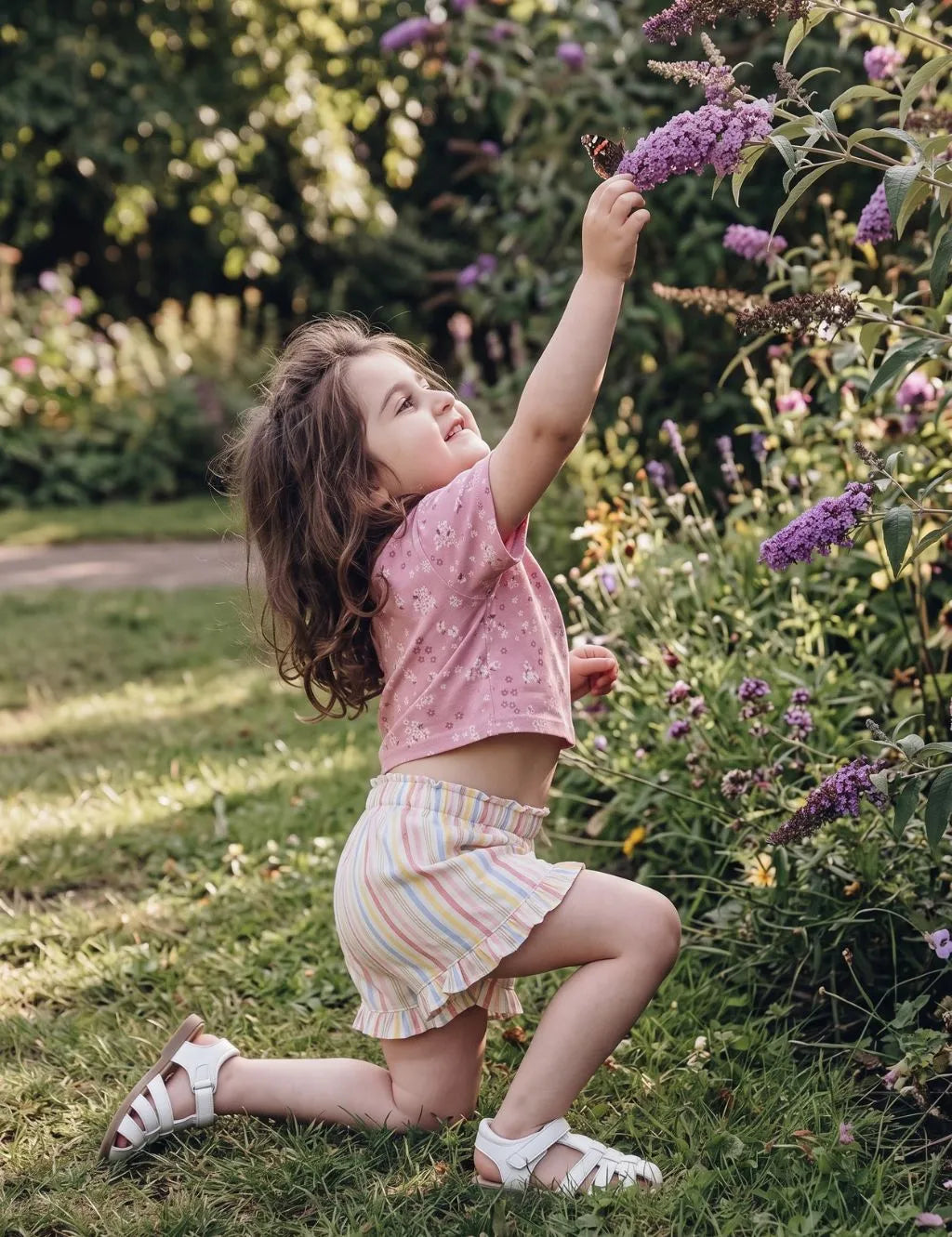 Little girl in an organic cotton pink floral shirt and pastel striped shorts kneeling in a garden reaching for a butterfly