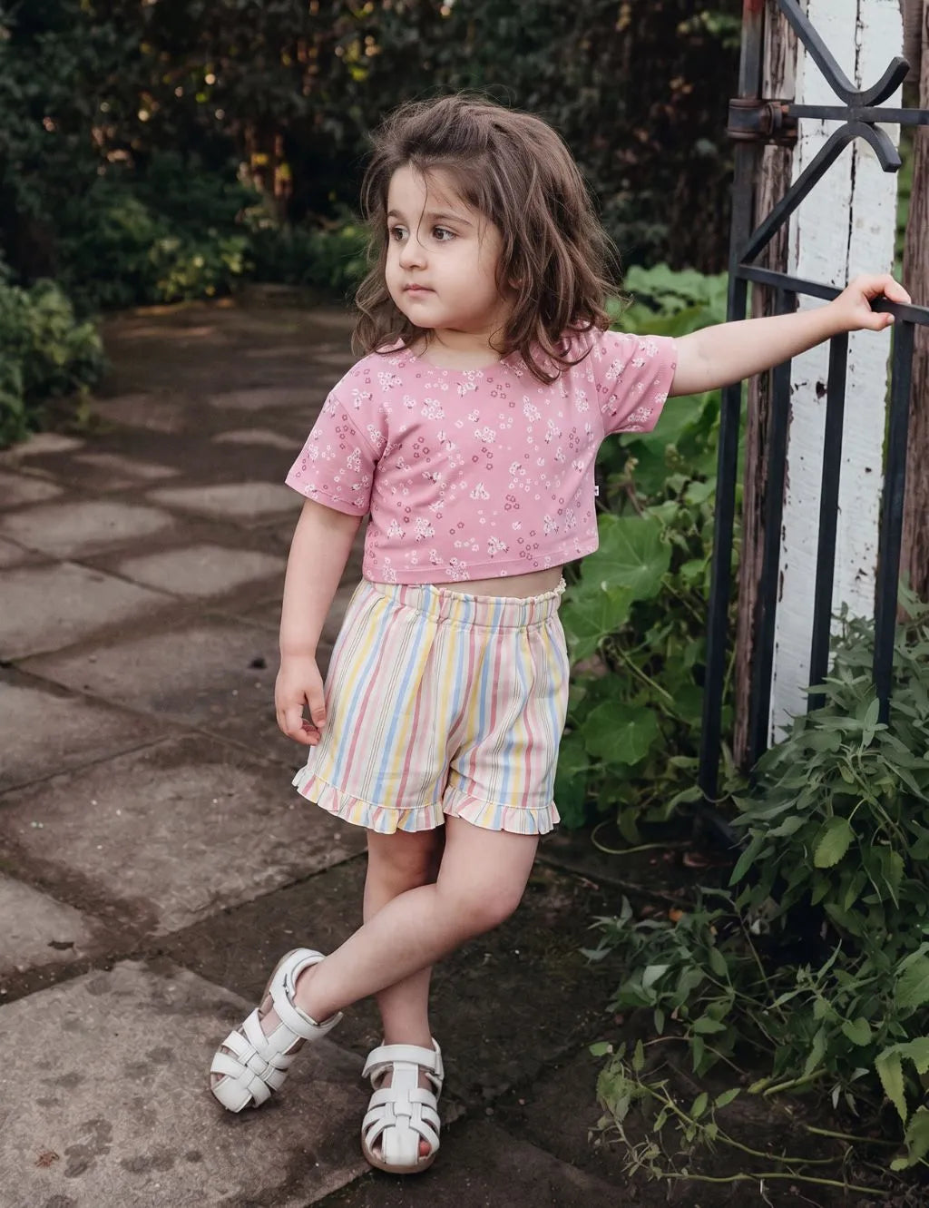 Little girl wearing an organic cotton pink floral t-shirt and pastel striped shorts standing outdoors near a gate.