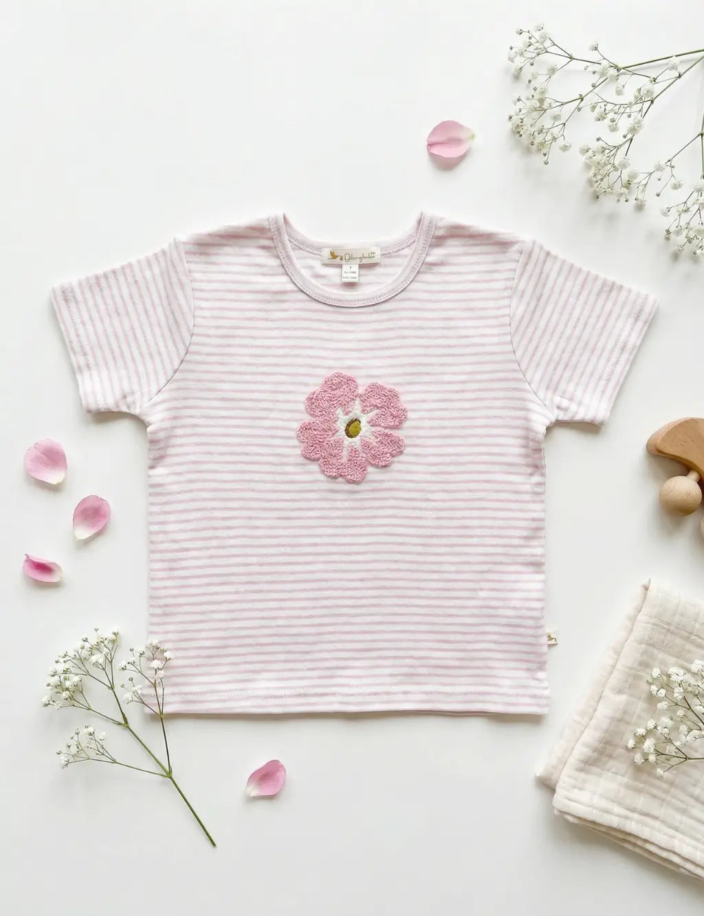 Flatlay of an organic cotton light pink striped short-sleeve top featuring a textured pink floral appliqué, styled with flower petals and baby's breath.