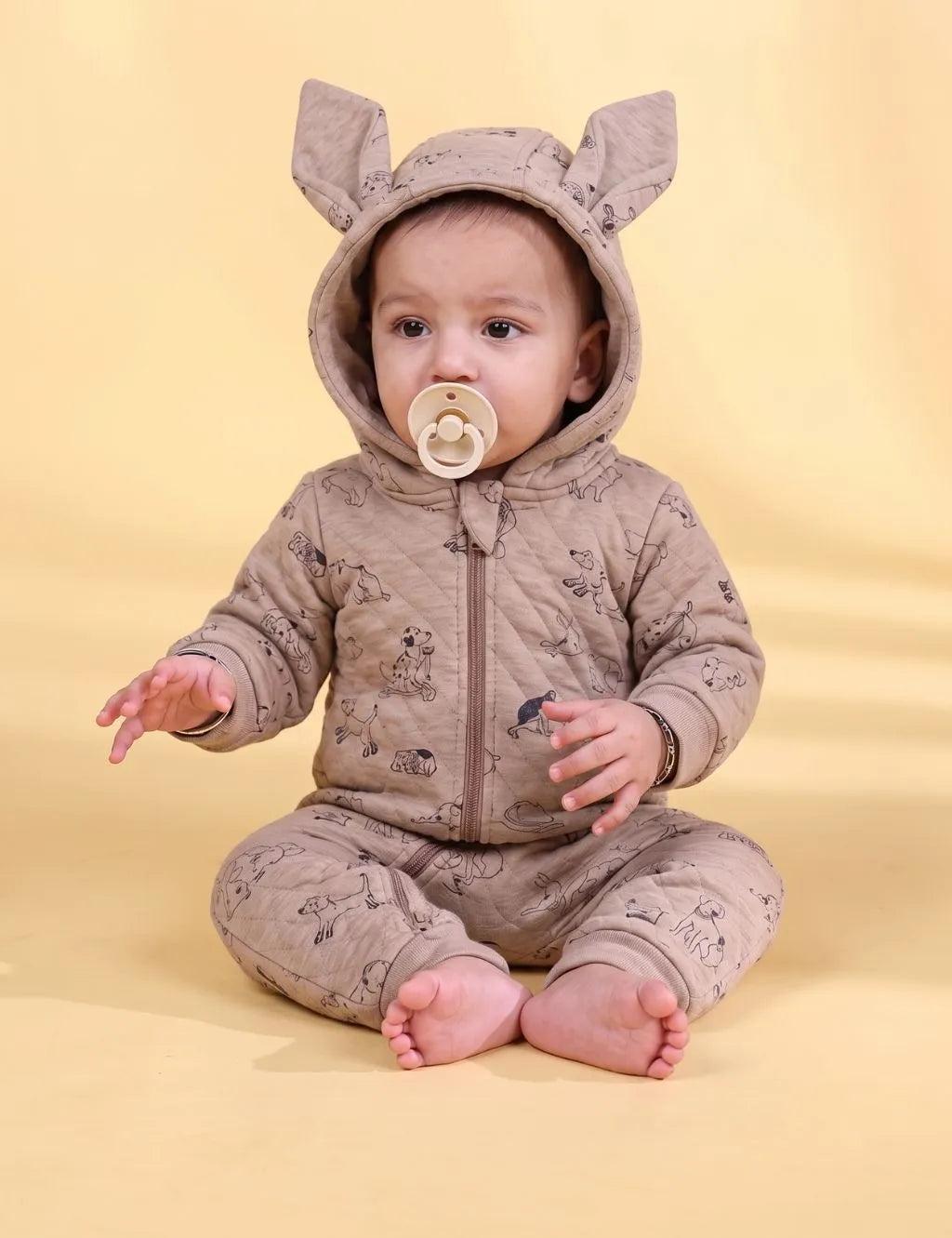 Baby sitting in a quilted beige hooded dog print romper with 3D animal ears holding a pacifier