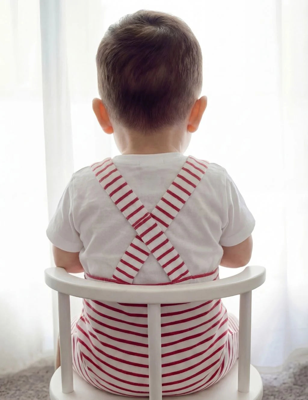 Back view of a toddler sitting on a white chair, wearing an organic cotton red and white striped onesie with criss-cross straps over a white t-shirt.