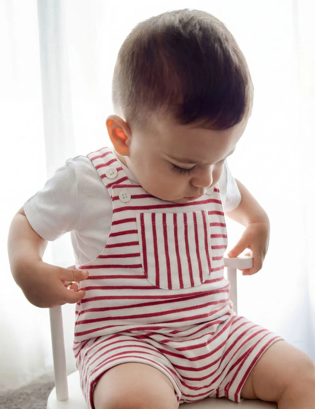 A toddler sitting in a white chair, wearing an organic cotton red and white striped onesie with a front pocket over a white t-shirt