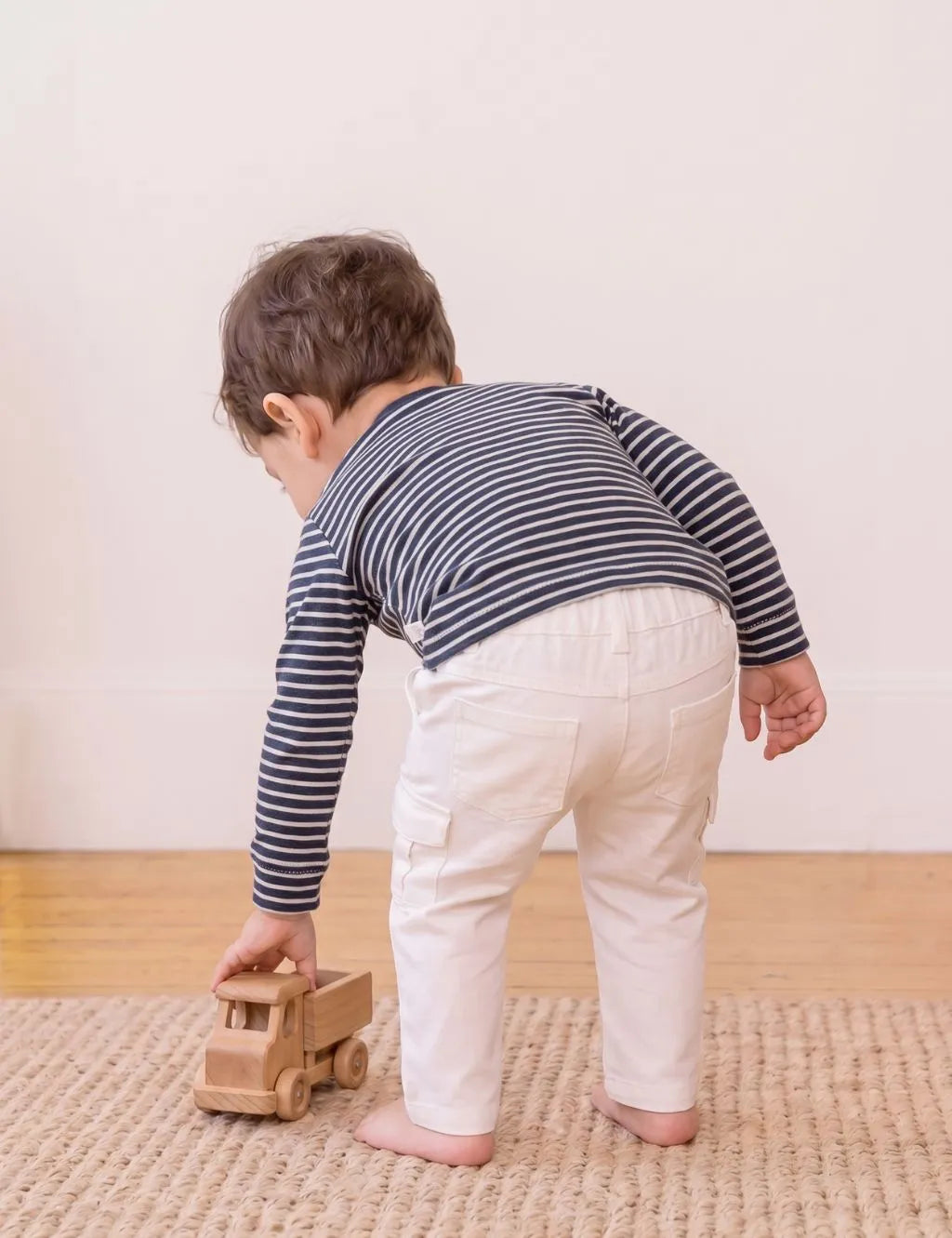 Back view of a toddler wearing an organic cotton striped polo shirt and white pants while playing with a wooden toy truck