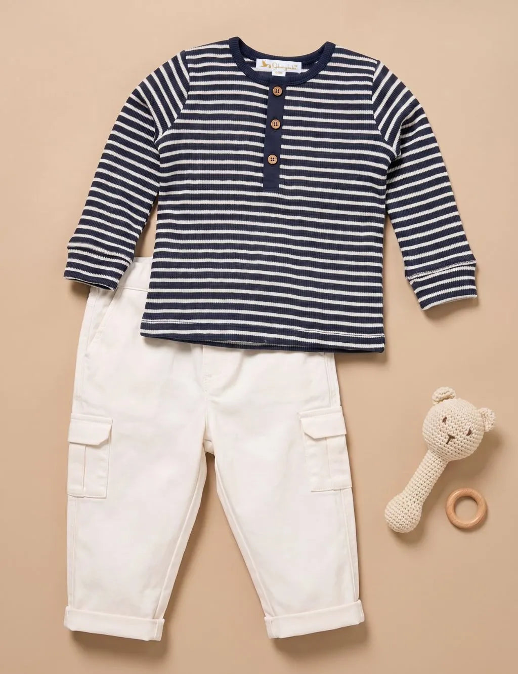 A baby clothing set featuring a navy and white striped long-sleeve henley shirt with wooden buttons, paired with off-white cargo pants, styled with a crocheted teddy bear rattle and wooden ring on a beige background.