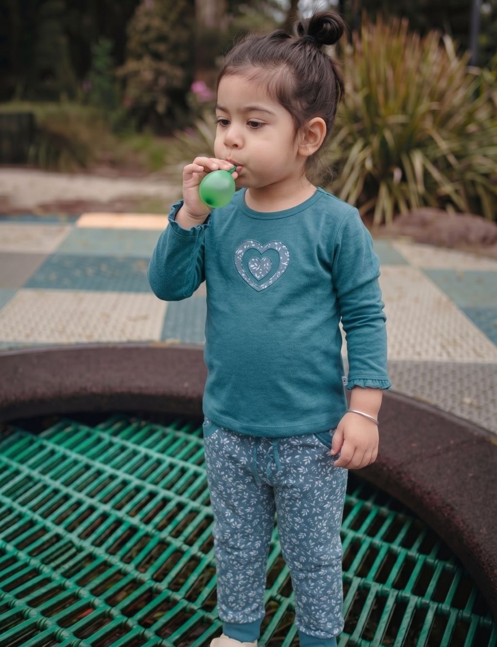 Little girl wearing an organic cotton teal long-sleeve shirt with heart embroidery and matching patterned pants outdoors.