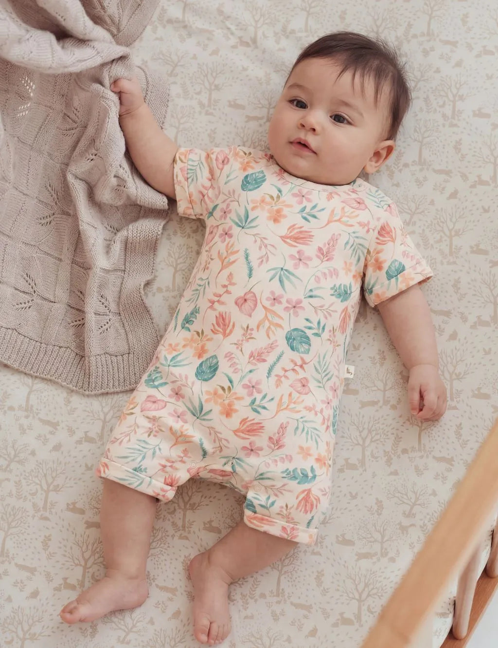 Baby lying down next to a knit blanket wearing a light-colored organic cotton short-sleeve baby romper featuring a vibrant peach and green Tropicana floral and leaf print.