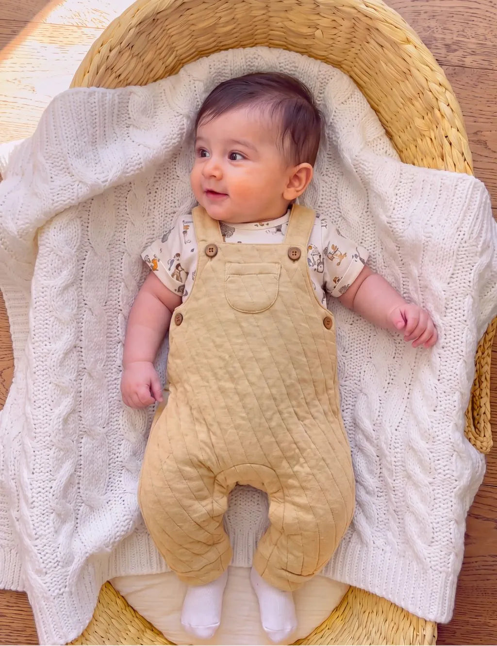 Baby lying in a wicker basket on a white cable knit blanket wearing an organic cotton patterned t-shirt and yellow quilted dungarees with white socks