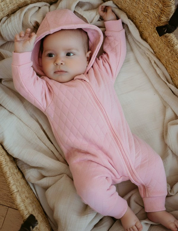  Newborn wearing organic quilted pink baby jumpsuit with hood, cozy full-body outfit for infants on a soft blanket.