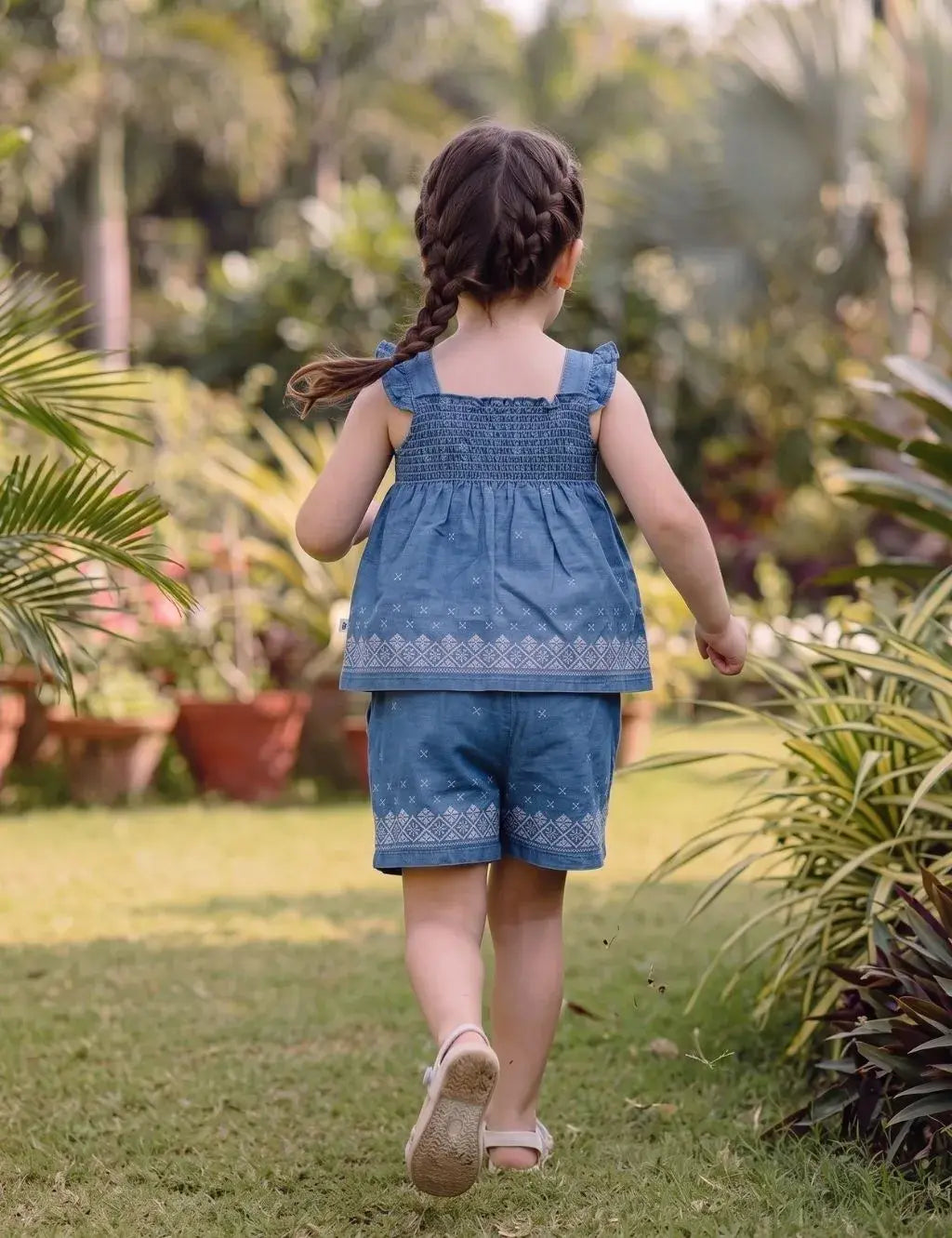 Back view of a young girl model with braided hair walking outdoors, wearing an organic cotton denim blue smocked top and shorts set with white embroidery.