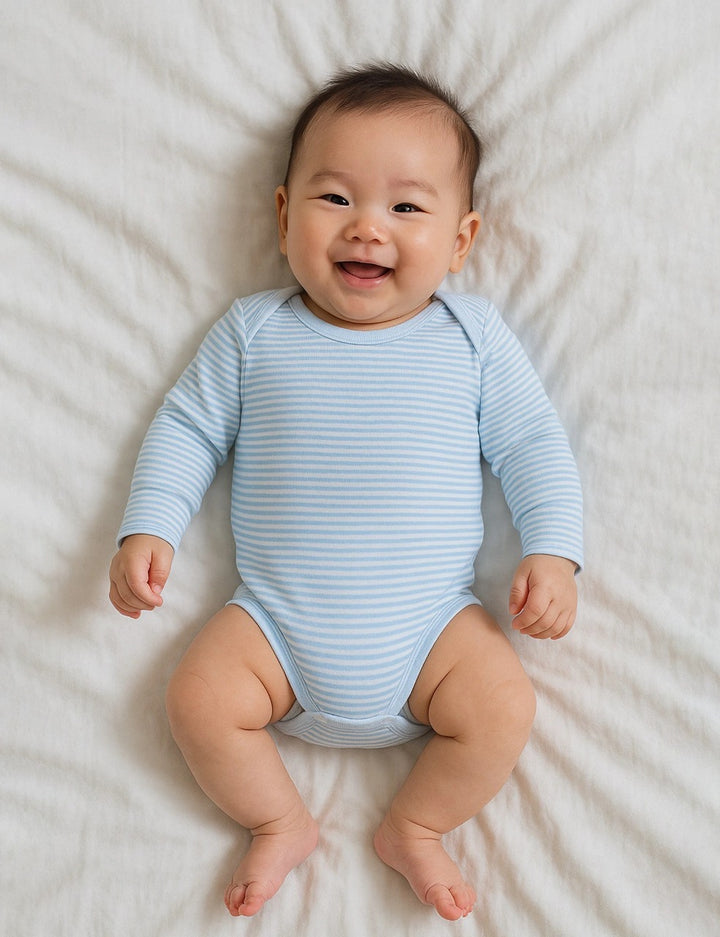 A happy baby, who appears to be of East Asian descent, lying on their back wearing a light blue striped organic cotton long-sleeve bodysuit. Shop comfy baby bodysuits India.