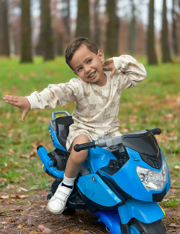 A cheerful child, who appears to be of Caucasian descent, sitting on a blue toy motorcycle wearing a light grey organic cotton sweatshirt and shorts with a festive bear print. Shop comfy baby outfits India.