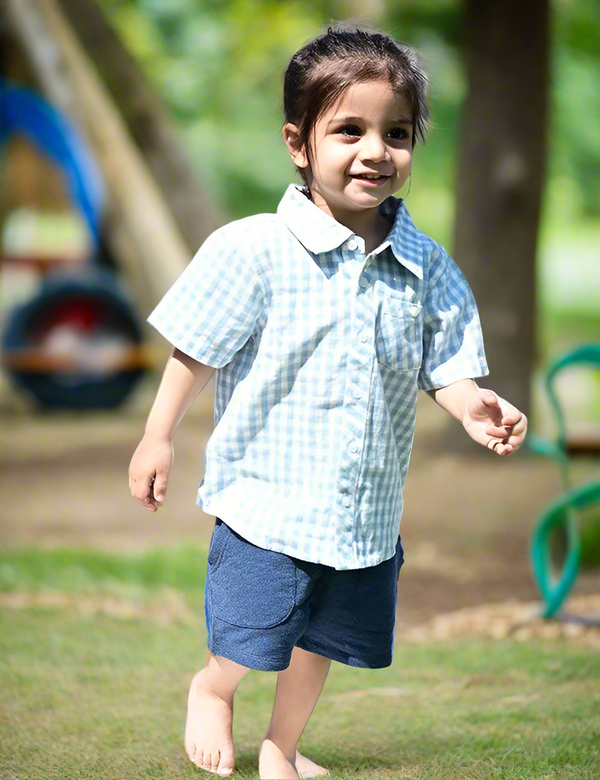 A young child, who appears to be of South Asian descent, standing barefoot and wearing a light blue and white gingham organic cotton shirt and navy blue shorts. Shop summer baby outfits India.