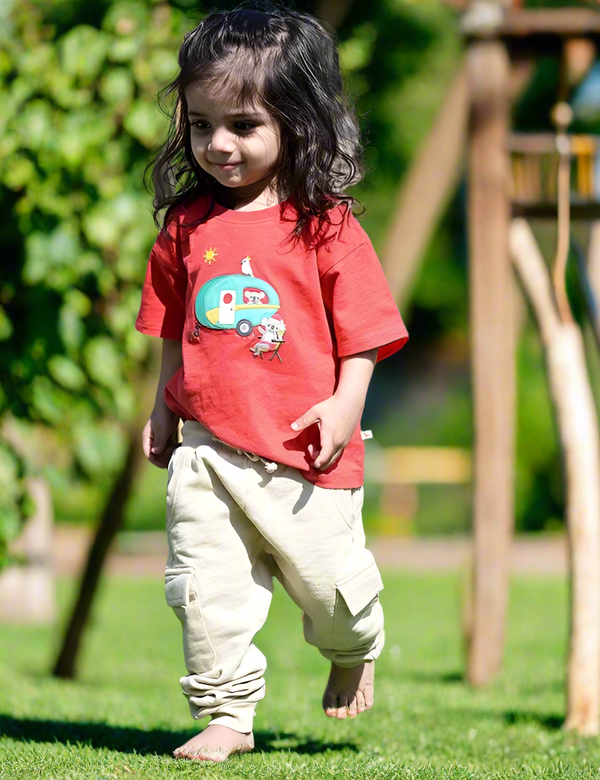 A young child, who appears to be of South Asian descent, standing barefoot and wearing a red organic cotton t-shirt with a koala camper van print and beige cargo pants. Shop cute baby outfits India.