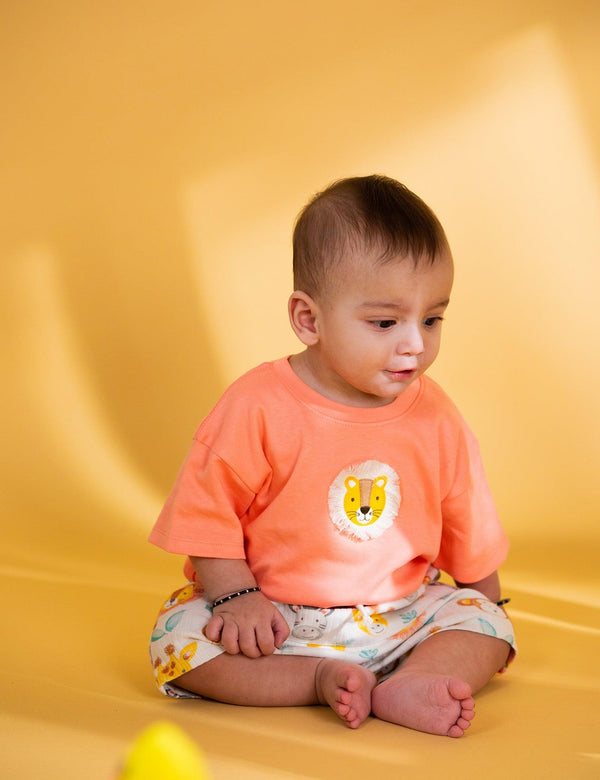  A cute baby, who appears to be of mixed ethnicity, sitting and looking down while wearing an orange organic cotton t-shirt with a lion patch and safari print shorts. Shop adorable baby outfits India.