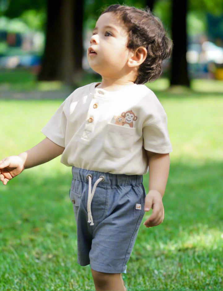  A cute child, who appears to be of South Asian descent, looking up while wearing a cream organic cotton henley shirt with a monkey pocket and blue shorts. Shop adorable baby outfits India.