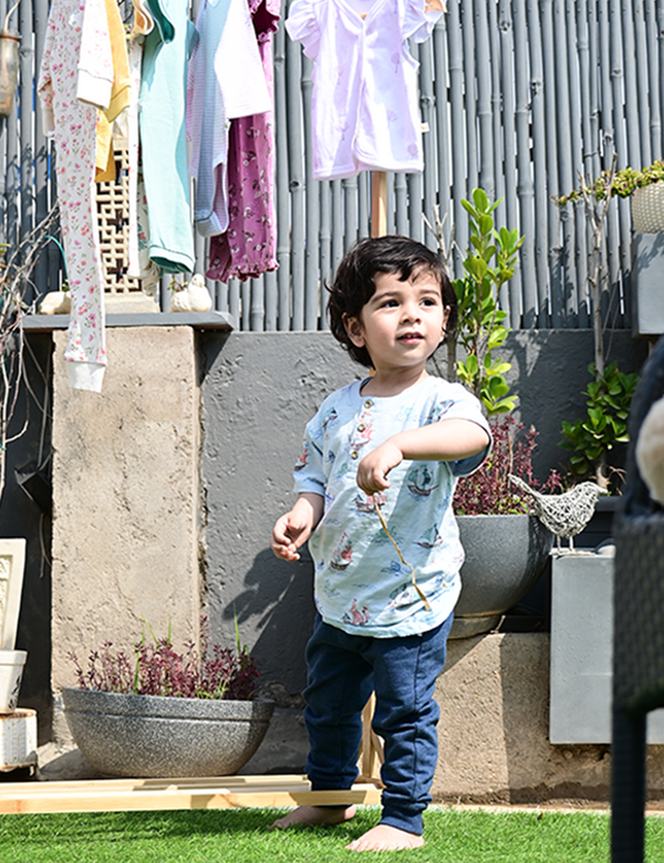 A young child, who appears to be of South Asian descent, standing barefoot and wearing a light blue sailboat print organic cotton t-shirt and navy blue joggers. Shop baby outfits India.