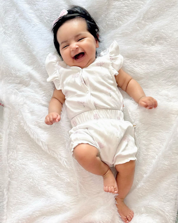 A joyful baby, who appears to be of South Asian descent, lying on a soft white blanket wearing a white organic cotton top with a winter tree print and matching bloomers. Shop cute baby outfits India.