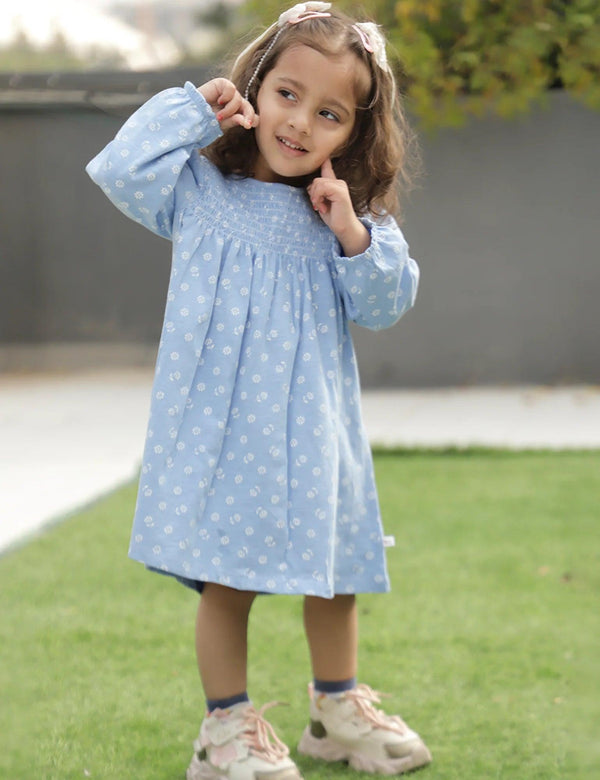A young child, who appears to be of South Asian descent, standing outdoors wearing a light blue organic cotton smocked dress with a ditsy print. Shop cute baby dresses India.