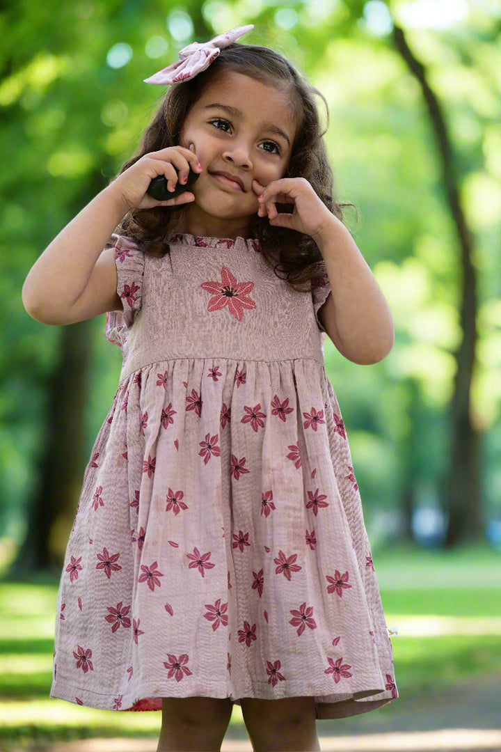A young child, who appears to be of South Asian descent, looking charming in a lilac floral muslin dress with a matching bow headband. Shop cute kids' fashion India.