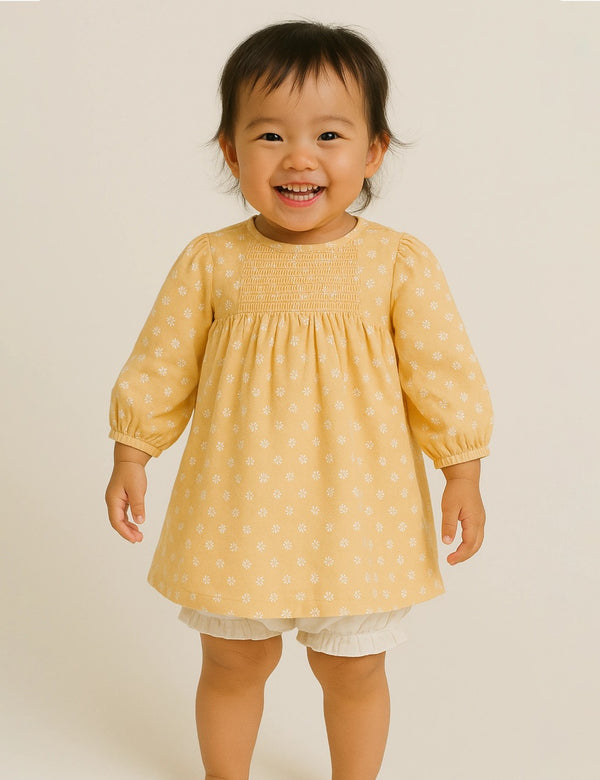 Adorable baby, who appears to be of East Asian descent, smiling while wearing a yellow organic cotton smocked dress with a white ditsy print. Shop cute baby dresses India.