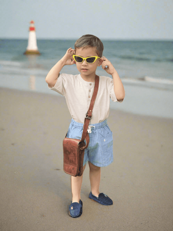 A young child, who appears to be of Caucasian descent, on a beach wearing a 100% organic cotton henley tee and blue striped shorts with sunglasses. Shop stylish baby sets India.
