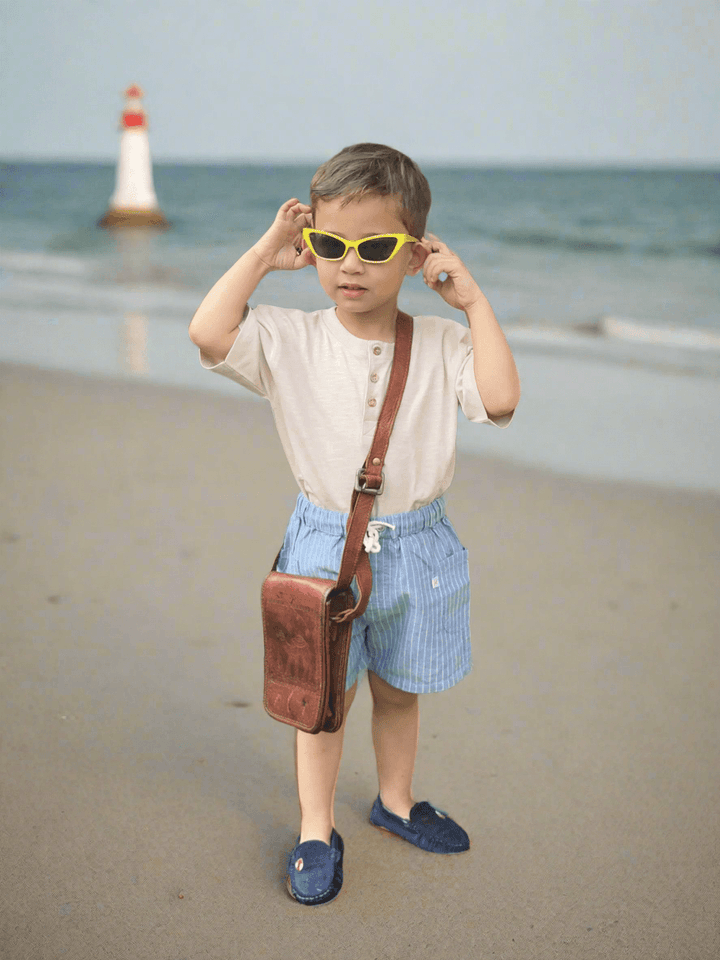 A young child, who appears to be of Caucasian descent, on a beach wearing a 100% organic cotton henley tee and blue striped shorts with sunglasses. Shop stylish baby sets India.