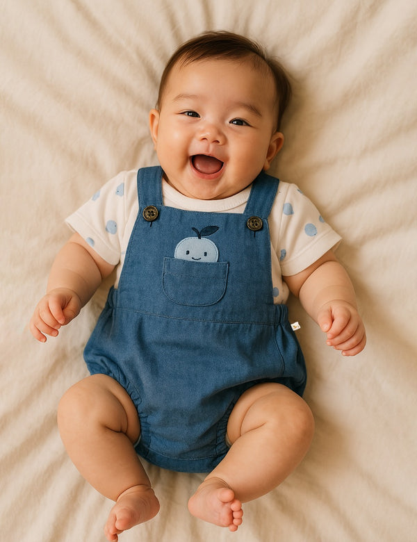 A happy baby, who appears to be of East Asian descent, lying on their back wearing a blue organic cotton dungaree with an apple pocket and a white apple print t-shirt. Shop cute baby clothes India.