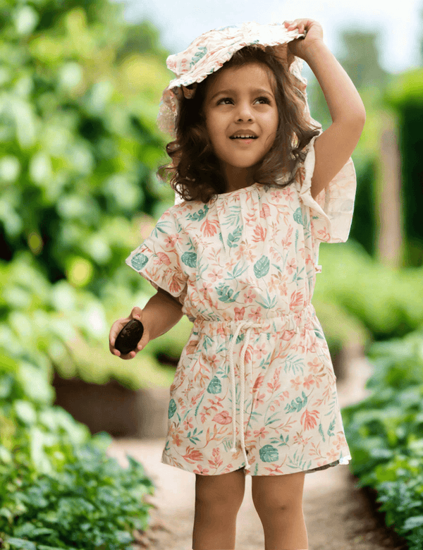 A happy young child, who appears to be of South Asian descent, outdoors wearing a floral print organic cotton romper and matching sun hat. Shop stylish baby outfits India.