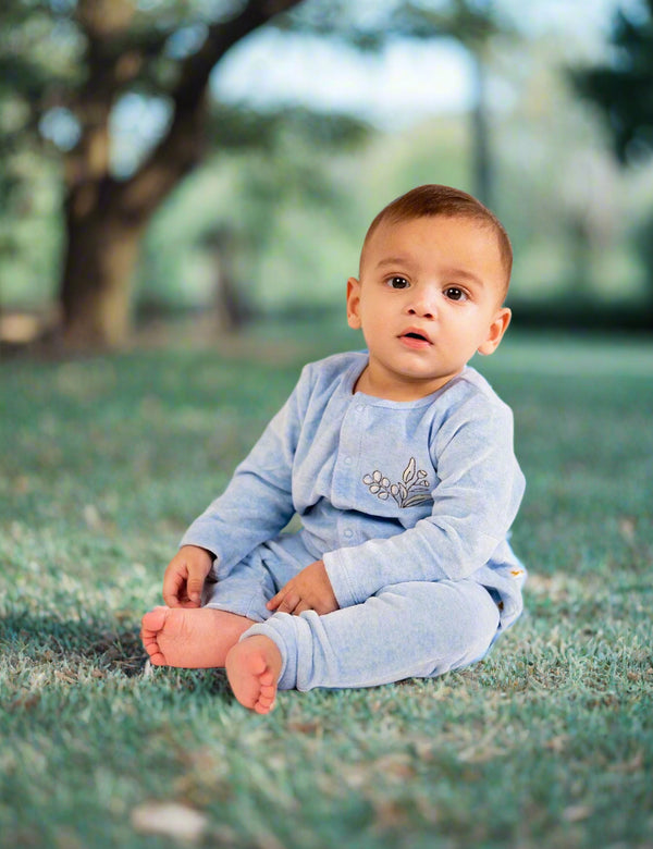 A serene baby, who appears to be of Middle Eastern descent, sitting on grass wearing a soft blue organic cotton cardigan and pants set with an olive branch embroidery. Shop comfortable baby outfits India.