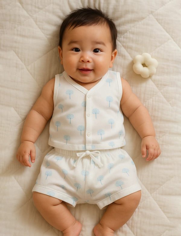 A happy baby, who appears to be of East Asian descent, lying on their back wearing a white organic cotton sleeveless top and shorts with a blue winter tree print. Shop cute baby clothes India.