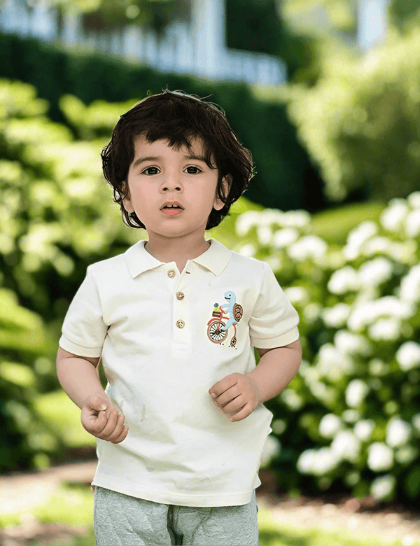 A young child, who appears to be of South Asian descent, standing outdoors wearing a white organic cotton polo with a turtle on a bike and brown pants. Shop cute baby outfits India.
