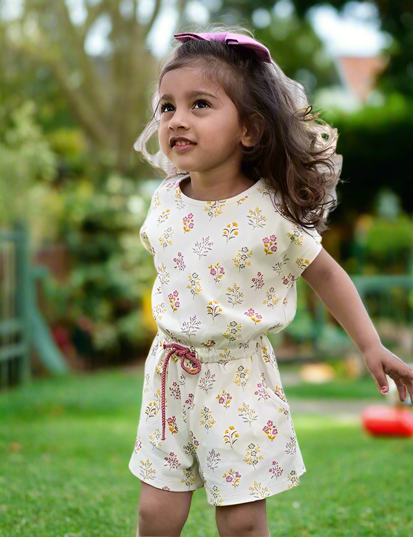 A young child, who appears to be of South Asian descent, playing outdoors in a white organic cotton romper with a delicate floral print and a mauve bow. Shop cute baby rompers India.