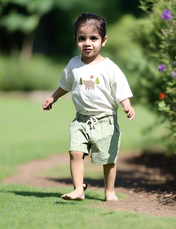 A sweet toddler, who appears to be of South Asian descent, walking barefoot wearing a bear appliqué organic cotton t-shirt and striped shorts set. Shop cute baby clothes India.